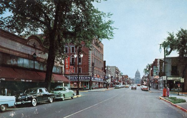 Color postcard of the State Street and Lake Street intersection, with the Co-op book store, the Rennebohm Drug Store and the Wisconsin State Capitol at the end of the street.