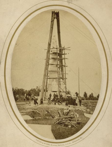 Governor Lucius Fairchild of Wisconsin and an unidentified group visiting the still uncompleted national monument at Gettysburg, Pennsylvania in 1869. Fairchild is just to the right of the monument, wearing a top hat. As the colonel of the 2nd Wisconsin Infantry, Fairchild was injured at the battle and his left arm was amputated. Also in the photograph is a portion of the National Soldiers' Cemetery and a group of soldiers in Zouave uniforms.