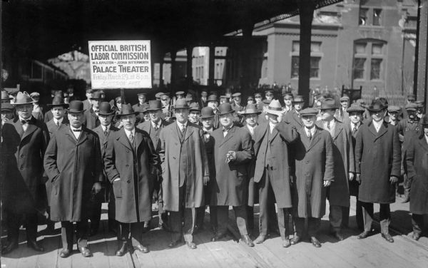 A large group of men in overcoats and hats posing together with a sign that reads: "Official British Labor Commission, W.A. Appleton — Joshua Butterworth, Palace Theater, Friday March 29 at 8 PM, Admission Free." William Rubin is in the front row at the far left-hand side.