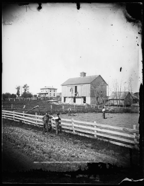 Two men are leaning on a fence in the foreground, with other men behind them in a field. Men are on scaffolding and ladders build a barn with a cupola. Other buildings include a log building on the side of the barn and an Italianate house in the background.