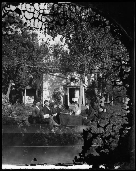 The Newhouse family sits around a table under trees with a book, goblets, a vase of flowers, a pitcher, a stereoscope and a cloth on the table in front of a frame house. Over the entrance to the house is a sign that reads "Hekla" indicating that the house is insured by the Hekla Fire Insurance Company. From left to right, they are: Christian Newhouse, Ole Newhouse, unknown, Knut Newhouse, Rachel Larson, Anna Newhouse, and Kari Newhouse.