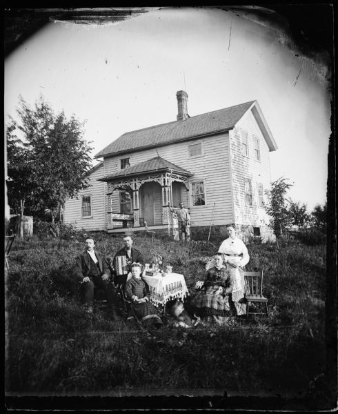 A man with an accordion, another man with a clay pipe, and three women sit around a table that has a knit cloth, a vase, a book, plants and fruit on it. Another man stands in the background by a two-story frame house. The house has carpenters lace on the front porch, cut work above the windows, a stone foundation and a lightning rod near the chimney.