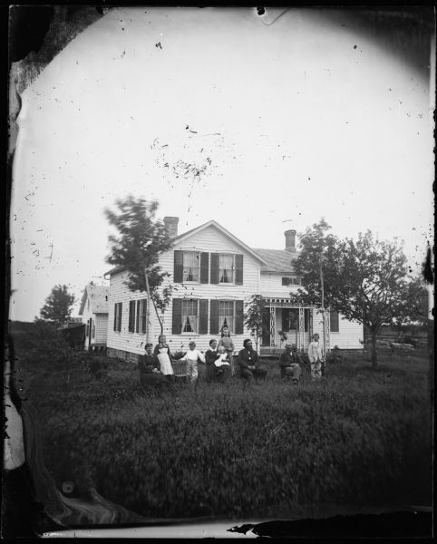 A family sits in the yard in front of their upright wing frame house. The house has cut work on the porch, small windows above the porch and a stone foundation.