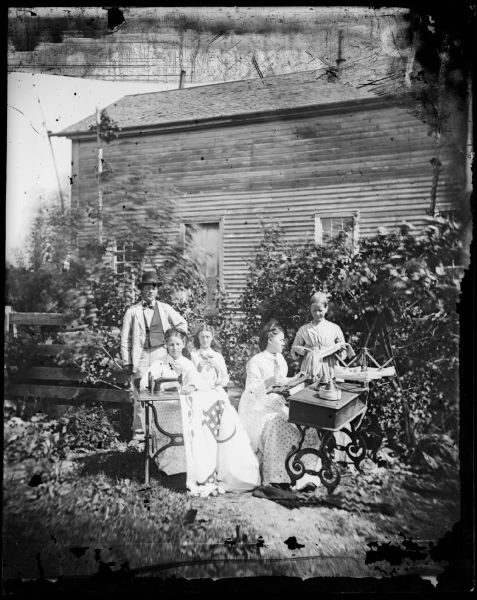 The G. Thompson family poses in front of their wooden house. One woman sits at a sewing machine working on a quilt while another works at a knitting machine.