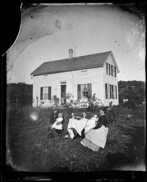 A couple and their young child and baby sit at a table in their yard. Behind them is a frame house with very small windows on the front second story and glass squares above the door. A sign leaning against the house reads: "Frihed Lighed Oplysning!" (Freedom and Equality in Education).