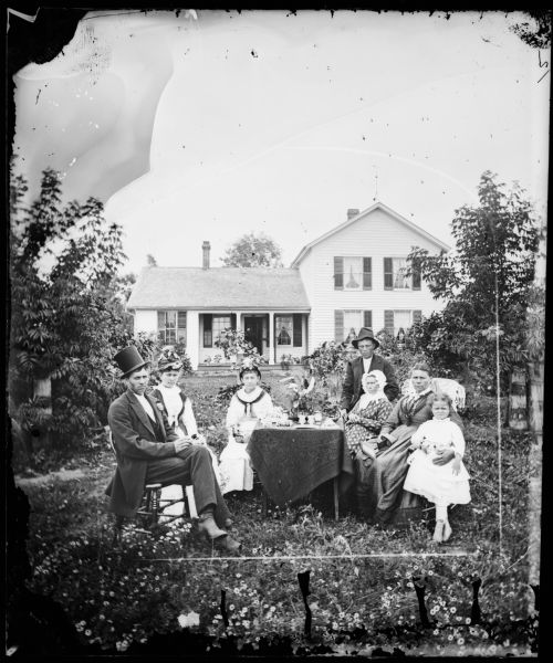 A family sits in a yard around a table with a cloth, vases and other objects on it. The man on the left is wearing a top hat and a flower in his lapel. The two women next to him are wearing decorated hats. The woman on the right is holding a stereoscope in her lap. Flowers are growing in the yard where the group is sitting. The family's upright and wing frame house is in the background.