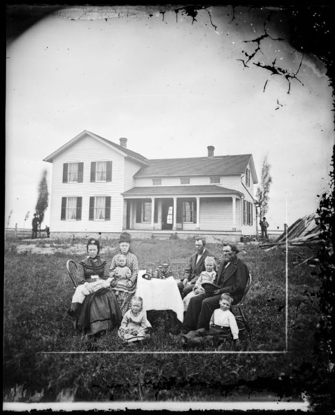 A family sits around a table with a stereoscope on it. An upright and wing frame house with a stone foundation is behind them. The woman has a large doll laid across her lap.
