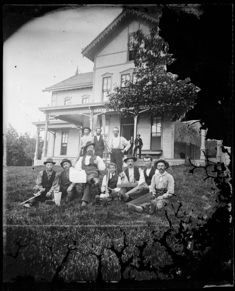 A group of men pose in the yard of the Tonyawatha House, later the Tonyawatha Spring Hotel with paddles and carpentry tools. The hotel is a frame bracket style structure that has latticework at the bottom of the porch and elongated windows overlooking Lake Monona. The hotel was under construction in 1879 and 1880.