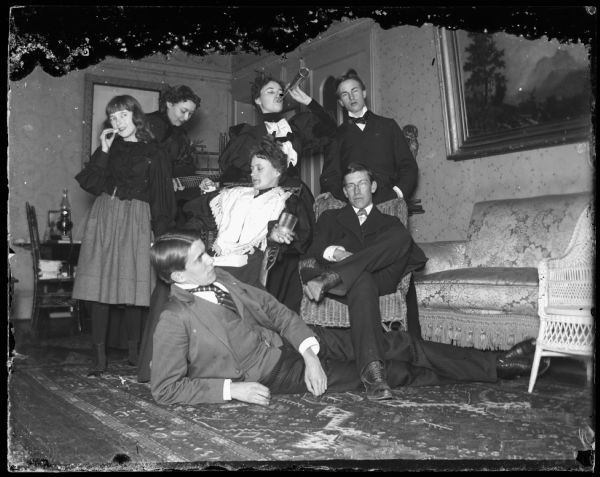 Group of people in dormitory or fraternity house. Four girls and three men inside room. One girl has a guitar and the other a small megaphone.