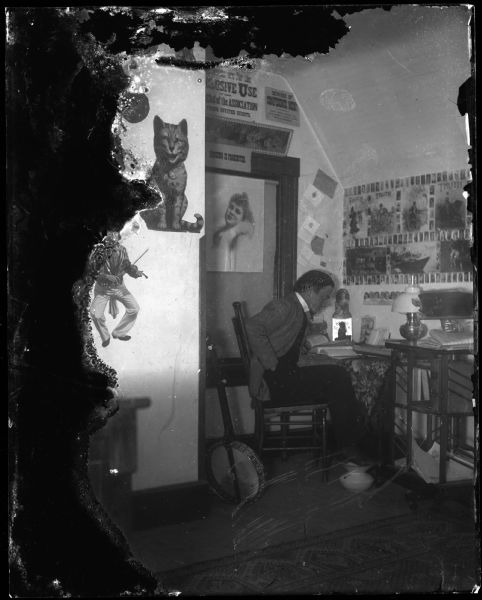 Man in dormitory room, with his feet resting on a spittoon. There is a banjo in the room and a picture of a cat on the wall.
