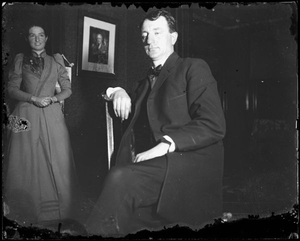 Informal portrait of seated man in a room with a woman in the background.