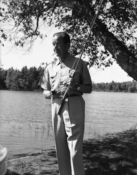 Edward R. Murrow stands with his fishing pole while smoking a cigarette on the shore of a lake in northern Wisconsin.