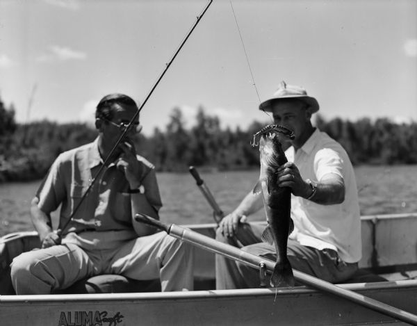 Edward R. Murrow sits in a boat smoking a cigarette, having just caught a Walleyed Pike while fishing in a lake in northern Wisconsin. His unidentified companion holds up the fish.