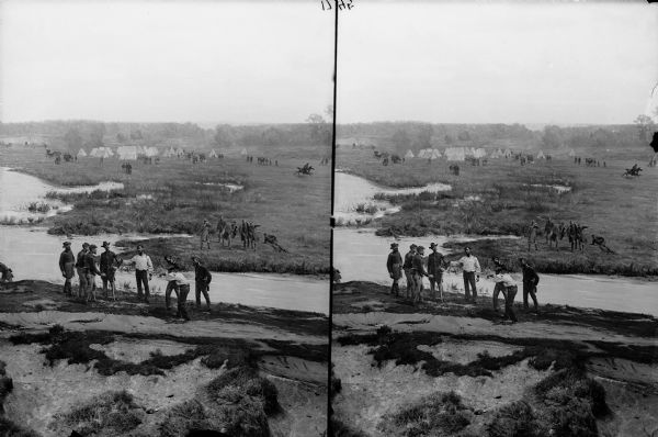Stereograph view of the Merrimac and Monitor Naval Battle cyclorama, painted by Théophile Poilpot. Men are standing on a riverbank in the foreground, and across the river are men in military uniform standing on the grass, with one man looking through binoculars. There are men around a camp in the background.