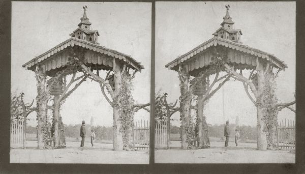Elaborate gateway, in the shape of an arch, to the National Soldiers Home, with man and boy standing in entrance. The gateway is made of trees, covered with a wooden roof, and including a miniature building on top.