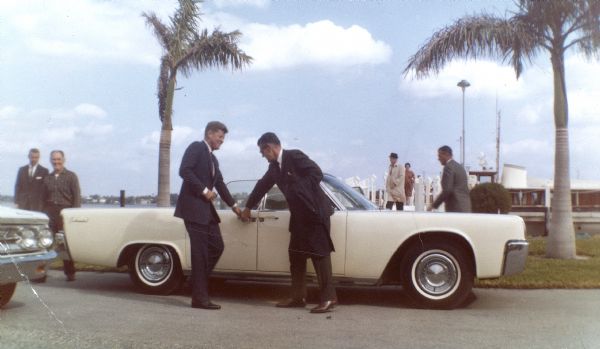 John F. Kennedy and another man by a white car at West Palm Beach. The caption on the back of the photograph reads, "on Lake Worth while visiting Fred and Selina". The man at left is U.S. Secret Service Agent Art Godfrey. The man opening the car door for the President is U.S. Secret Service Agent Gerry Behn. The man at right is U.S. Secret Service Agent Bill Greer.