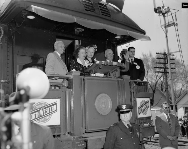 President Harry S. Truman greets a crowd from the back platform of a NorthWestern train at the Commercial Ave. crossing.  Others with him from the left are Governor Oscar Rennebohm, Mrs. Truman, and Mrs. Rennebohm.  He came for a 5 1/2 hour visit, gave a "peace" address to the nation from the UW field House and dedicated the new CUNA building on Sherman Ave. The man in dark glasses standing at the bottom right is U.S. Secret Service officer Floyd Boring.