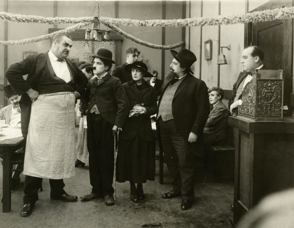 The huge waiter (played by Eric Campbell) glowers at the immigrant (Charlie Chaplin). To the right are Edna Purviance playing another immigrant and Henry Bergman as a bearded artist.