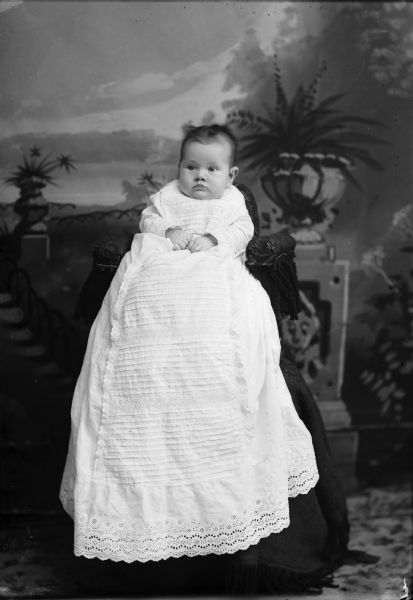Studio portrait in front of a painted backdrop of an unidentified European American infant sitting up on a high chair. The infant is wearing a long christening gown.