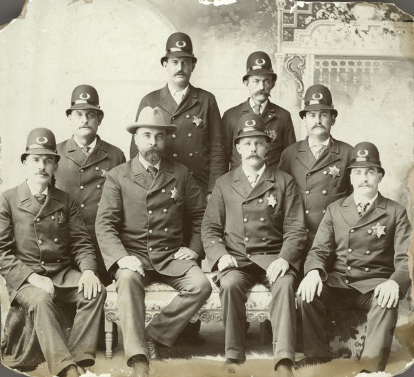Group portrait of Police Chief John Adamson and police officers. In the front row from the left are Frank Currier, John Adamson, Jake Togsland, and John Scherer. In the back row from the left are Tom Shaughnessy, Jake Behrend, Arne K____ (writing on print hard to read), and Alex O'Neill.