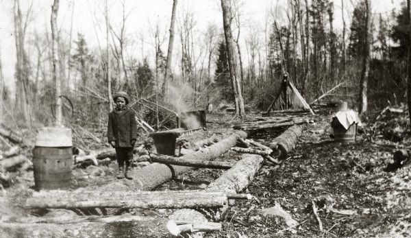 A child is standing on a log lying on the ground in a clearing. There is a barrel on the left, and a campfire in the background.