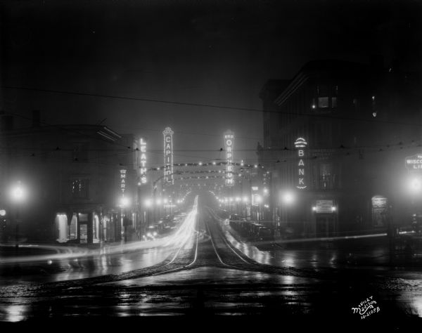 Night view looking down State Street from the intersection at North Carroll Street. Lighting up the scene are the blurred lights of automobiles moving along the street, Christmas lights arched over the street, and the illuminated marquees and signs of the Capitol and Orpheum Theatres, Leath's Furniture, the Commercial National Bank, and Mark Martin's restaurant.
