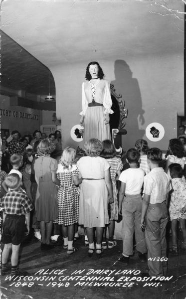 A crowd surrounds a 10-foot mechanical mannequin of Alice in Dairyland at an Alice display for the Wisconsin Centennial Exposition. The Alice could raise her arms, turn her head and sit down on her over sized throne. Caption reads: "Alice in Dairyland, Wisconsin Centennial Exposition, 1848-1948 Milwaukee, Wis."