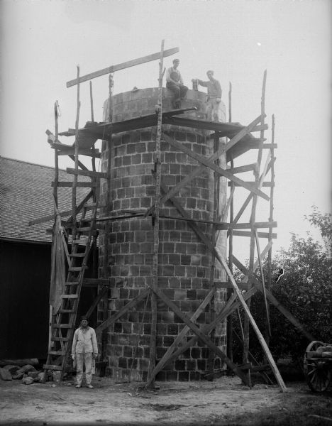 Three men pose next to a silo that is under construction, with wood scaffolding around it.