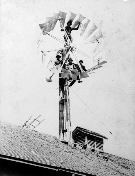 Several men, including Frank Klug, pose on the blades of a windmill positioned on the roof of a building.