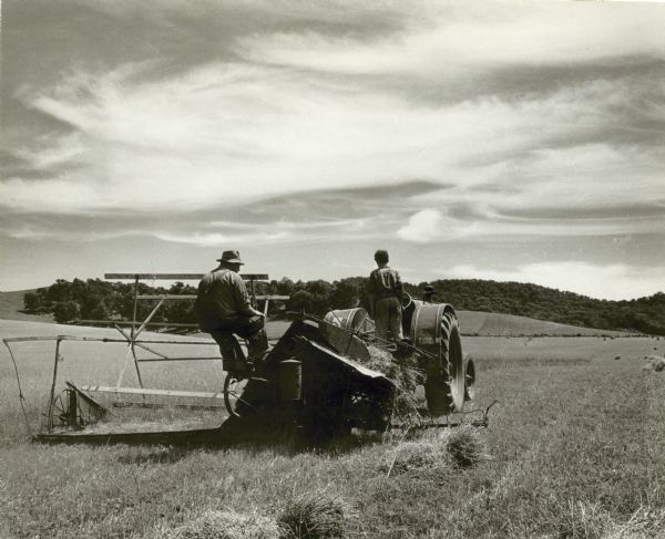 A farmer and a young boy ride a tractor through a field reaping oats.