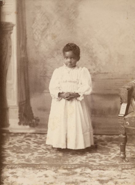 Studio portrait in front of a painted backdrop of an African American child (probably a girl) wearing a white dress. Text written on the back of the photograph reads "Mrs. Butts, next week, middle, 633 E. Johnson," which is probably a photographer's note.
