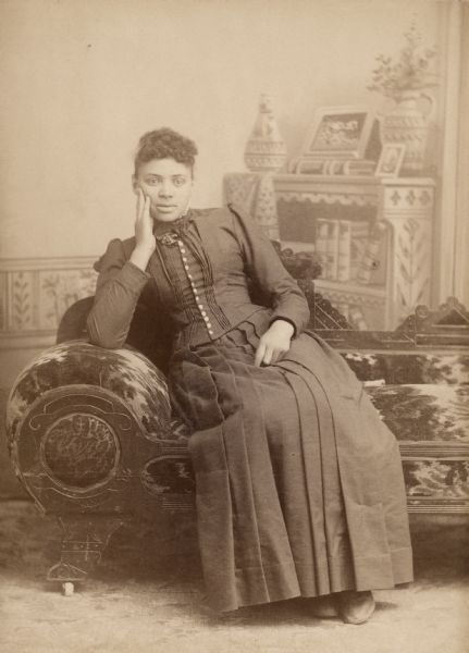 Studio portrait in front of a painted backdrop of an African American woman in a formal dress seated on a sofa. Photograph was taken by Berdette E. Akin, who was an active photographer in Hillsboro from 1887-1908.