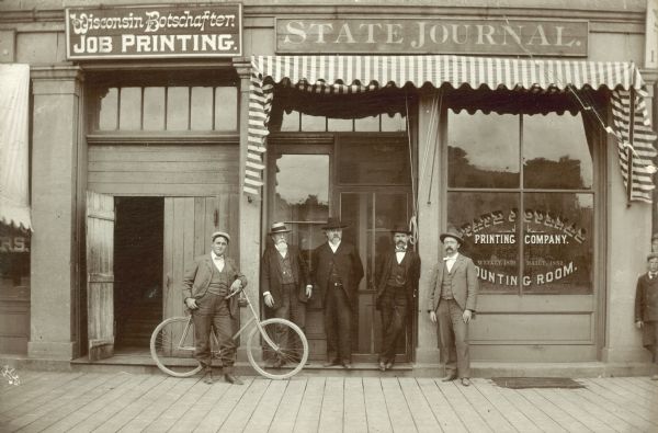 Exterior view of the offices of the "Wisconsin State Journal" at 117 East Washington Avenue. Posing in front of the building are David Atwood, John Hawks, Fred Arthur and Walter Balley, and one unidentified man. One of the men is standing with a bicycle.