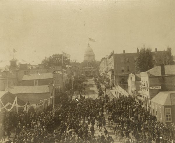 Elevated view of parade celebrating President Grover Cleveland's visit, coming up King Street to the Wisconsin State Capitol from the East Madison Depot. The Schlimgen Marble Works, visible at left center, was then located at the corner of King and Butler Streets.