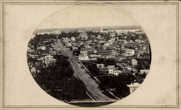 View up State Street looking toward the Wisconsin State Capitol from the roof of University Hall (later Bascom Hall), about 1860 or 1861.  Actually the photograph captures two Capitols: the first Capitol is the facade facing State Street, while the new, larger building then under construction can be seen behind it. Bruen's Block (built 1853) is to the left of the Capitol; to the right are the Vilas House, (built 1853), Grace Episcopal Church (completed 1858), First Baptist Church (dedicated 1854), Wells' Block , and the Dane County Courthouse. Lake Monona is visible at the top of the image.