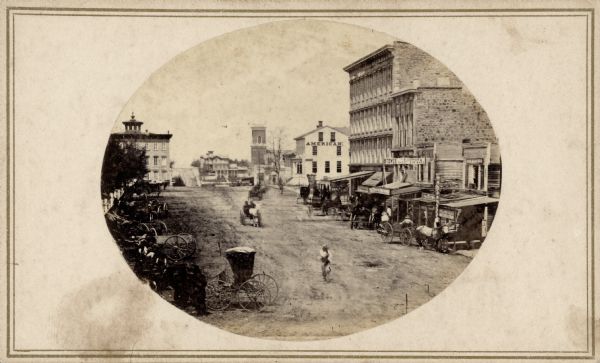 View of Pinckney Street, about 1860, from the roof of the Fairchild Block at the corner of East Main and South Pinckney looking northwest toward Lake Mendota. Bruen's Block is the large commercial building on the right. Next to it, across the East Washington intersection, is the historic American House hotel where the Legislature first met. On the left numerous horse-drawn buggies and wagons are hitched along Capitol Park fence. Bacon's Block is the building with the turret on its roof at the corner of Pinckney and Mifflin streets.