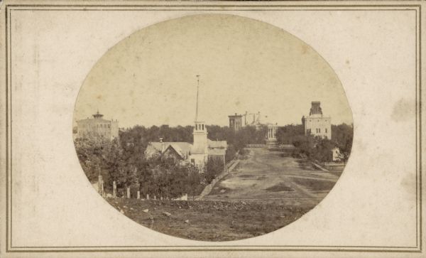 A view looking south down Wisconsin Avenue toward the Capitol Square where portions of the second and third Wisconsin State Capitols are visible. This perspective provides a better view of the odd appearance that the two Wisconsin Capitols made at the time, with the old Capitol, on the right, facing State Street, and the East Wing of the larger, new Capitol behind it, facing King Street. Other prominent buildings include, from the left, Bacon's Block, the Presbyterian Church, and on the right Madison City Hall at the corner of Wisconsin Avenue and West Mifflin Street.