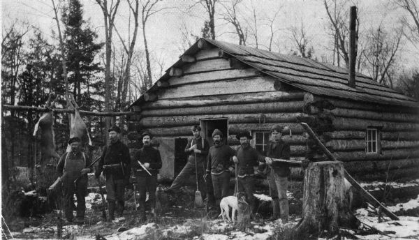 A group of men stand with rifles at the Echo Lake Hunting Club Camp.  Gutted deer hang to cure outside the cabin.