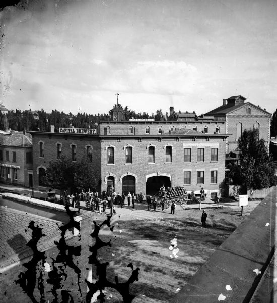 Elevated view of employees, along with young children, posed around a wagon loaded with barrels of beer in front of Hausmann's Capital Brewery at the corner of State and Gorham Streets. In the 1870s, Capital Brewery was the largest of five German owned and run Madison breweries. A group of men and young boys are standing on the roof near a sign that reads: "Malt-House."