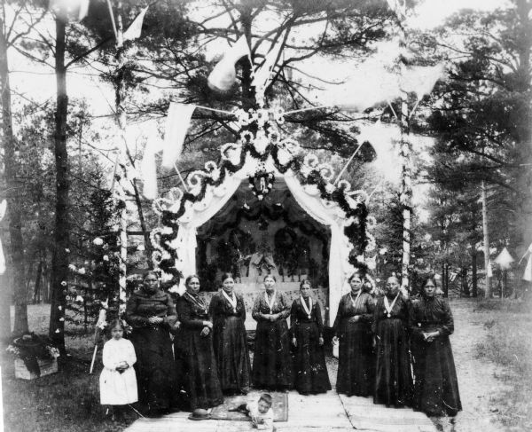 Members of the Menominee tribe pose during the Corpus Christi celebration, possibly held at the Keshena chapel. Pictured are, from left to right, Clara Chickney, Elizabeth Kakkak, Victoria Lafrombois, Teresa O'Katchicum, Agnes Prickett, Elizabeth Burnette, Mary Lookaround, Rose Be(?), and Elizabeth Shawanopenass. The boy in the center is Louis Duquaine.