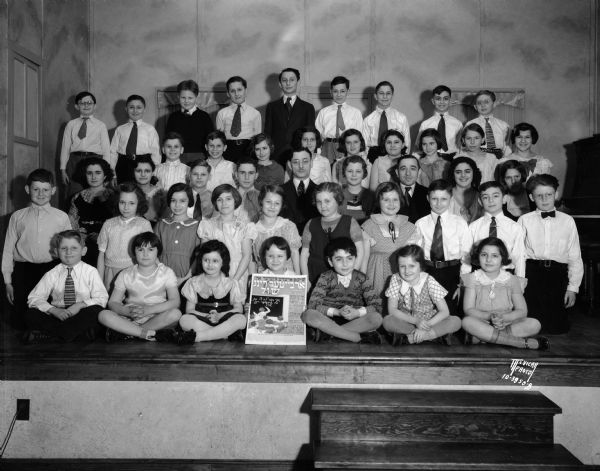 Large group of Jewish children sitting on stage at the Workmen's Circle Labor Lyceum, 41 N. Mills Street, in the Greenbush neighborhood. One child sitting in the front row is holding a sign with Hebrew writing. Two men and a woman are posing in the center row.