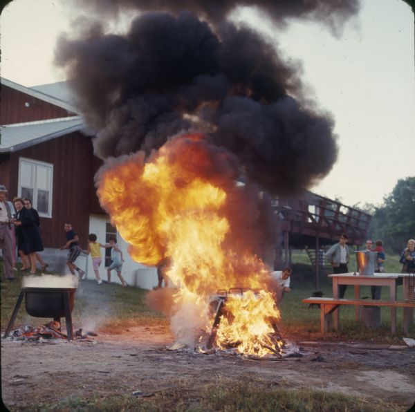 Outdoor view of two pots used for a fish boil. One pot is engulfed in flames and children are running from the area.