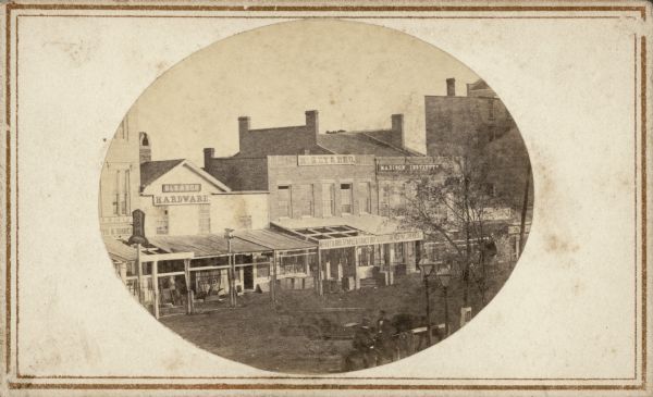 John S. Fuller took this photographs of the buildings in the first block of East Main Street from the Wisconsin State Capitol roof. Most of the businesses in the picture are housed in wooden, false-front structures. They include Gleason Hardware and the Madison Institute. Several men on horseback and two Capitol Park gas lights can be seen in the foreground.