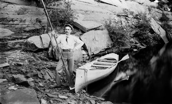 A river guide (possibly Clarence C. Bennett) holding an oar while posing next to his rowboat on shore.