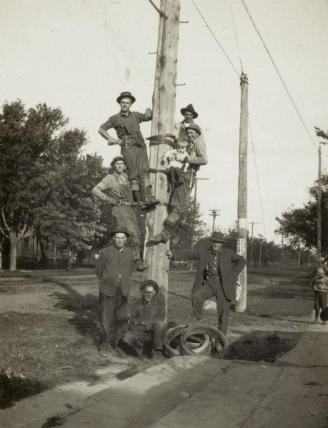 Several men in safety harnesses pose on and around a utility pole. One of them is holding a small child.