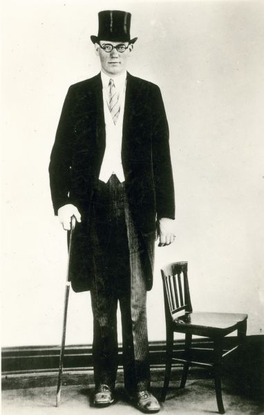 Portrait of Clifford Thompson, at one time the tallest man in America, in a jacket and top hat posing next to a chair.