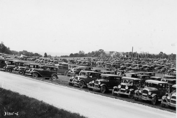 A large numbers of cars are shown parked at Peninsula State Park during the reburial ceremony for Potawatomi Chief Simon Onanguisse Kahquados. A crowd of people is beyond the parked cars near a totem pole. There are U.S. Flags flying.