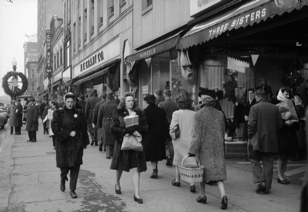 Christmas shoppers on South Pinckney Street on the Capitol Square.  Businesses include the Three Sisters store, Mangels store, W.T. Grant store and a sign for a Chop Suey restaurant.