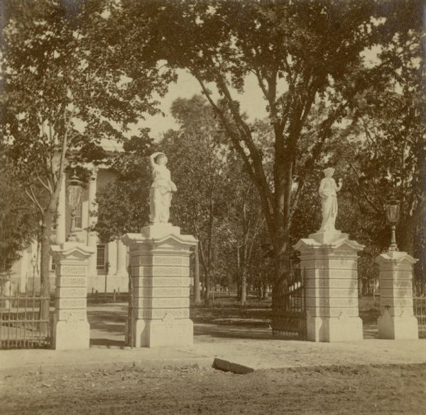 Southeast Gate of Capitol Park. Four stone columns stand along the sidewalk, with statues on top of the two columns in the center, and two columns with streetlamps flanking them on the left and right. In the background are trees and the Wisconsin State Capitol. The street in the foreground is dirt.