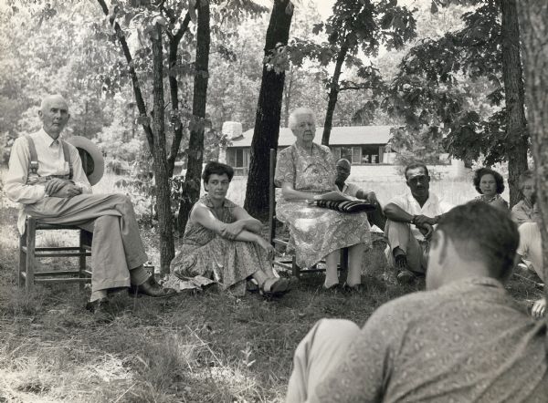 A group of people sitting on the grass and chairs outdoors for a meeting with the Highlander Library in the background. From left to right: Father of Myles Horton; Julie Mabee from the local community; Dr. Lillian Johnson; Mr. Paul Bennett; Mother of Myles Horton.
